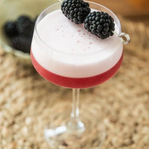 A pink cocktail with a frothy top is served in a coupe glass, garnished with two blackberries on a cocktail pick. The glass sits on a woven mat with a bowl of blackberries in the background.
