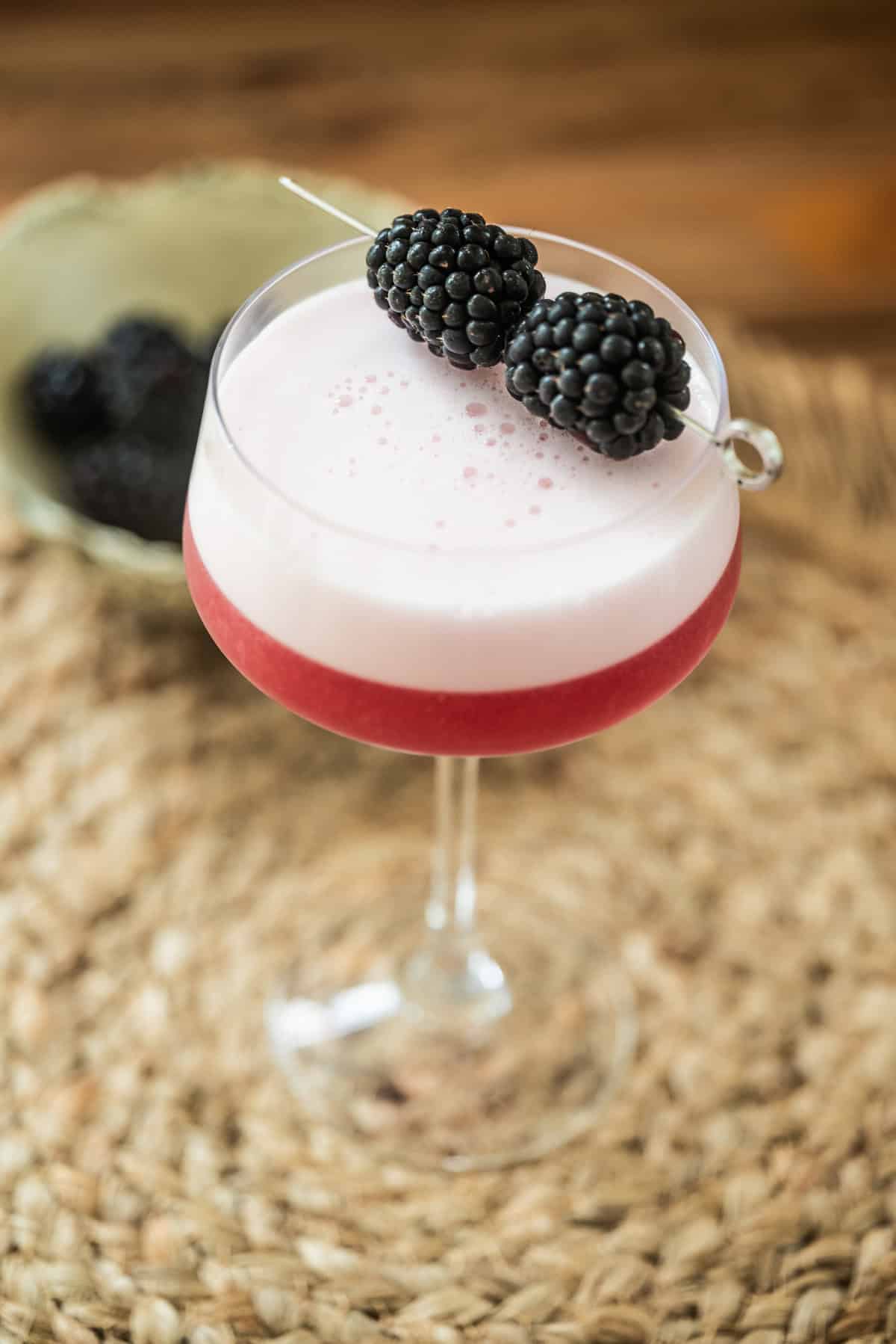 A pink cocktail with a frothy top is served in a coupe glass, garnished with two blackberries on a cocktail pick. The glass sits on a woven mat with a bowl of blackberries in the background.