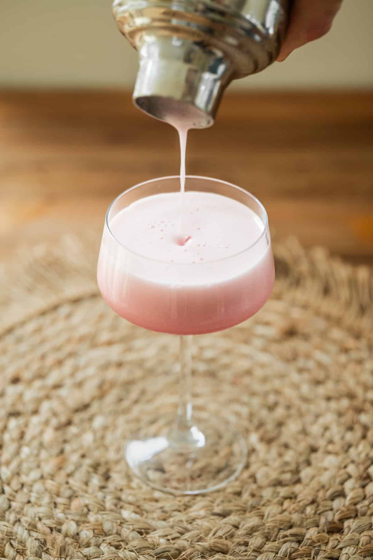 A creamy pink cocktail is being poured from a shaker into a stemmed glass, set on a woven placemat with a blurred wooden background.