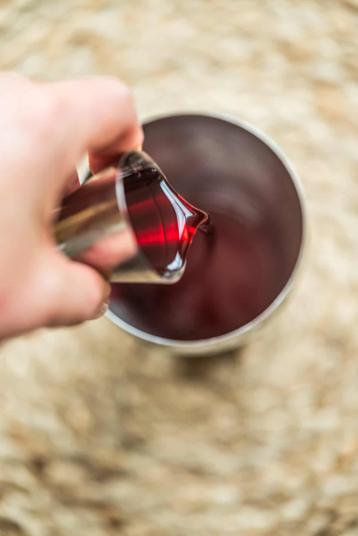 A hand pours a red liquid from a metal jigger into a metal cup, with a woven surface in the background.