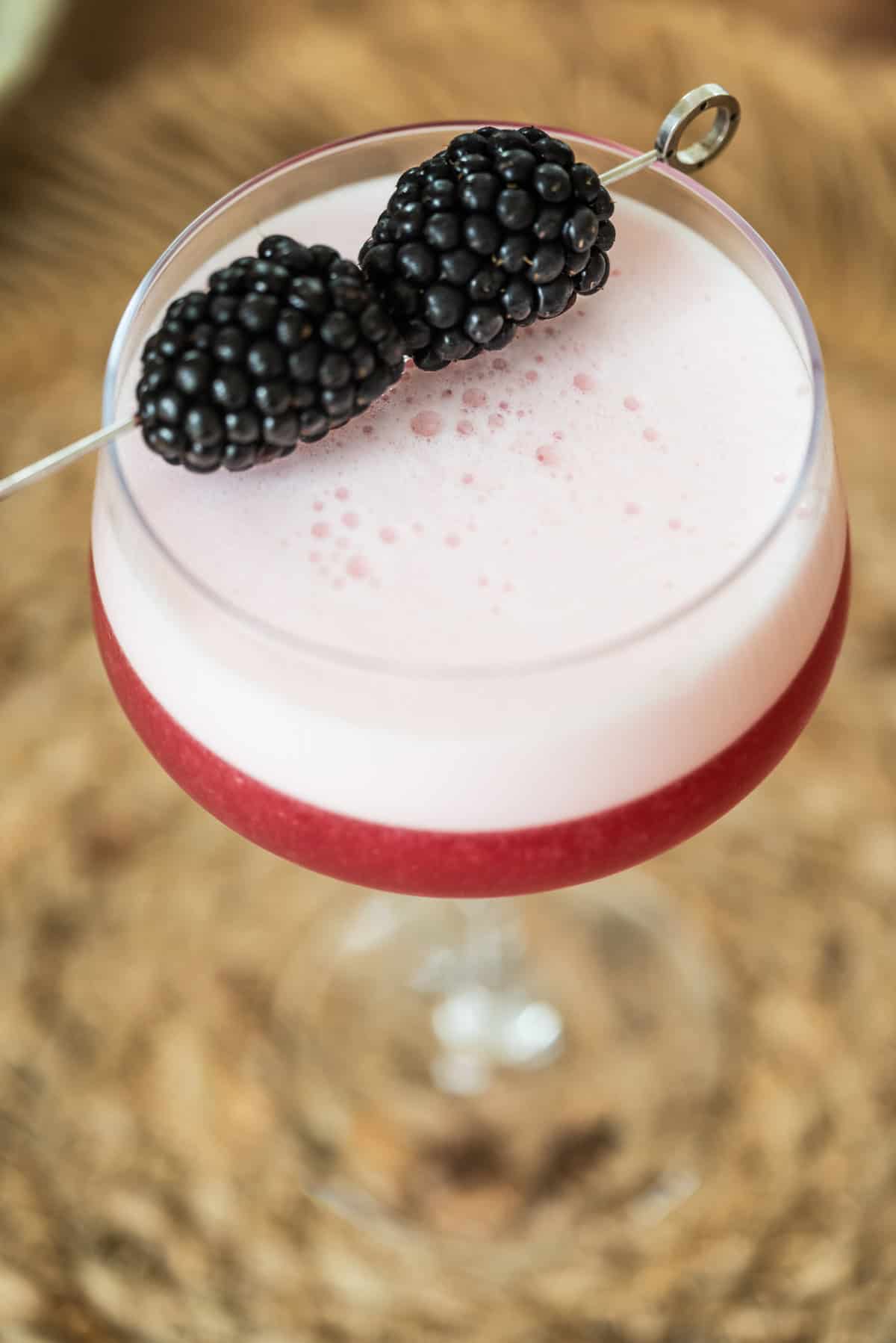 A close-up of a cocktail in a glass with a frothy pink layer on top and a deep red layer below, garnished with two blackberries on a cocktail pick. The background is blurred.