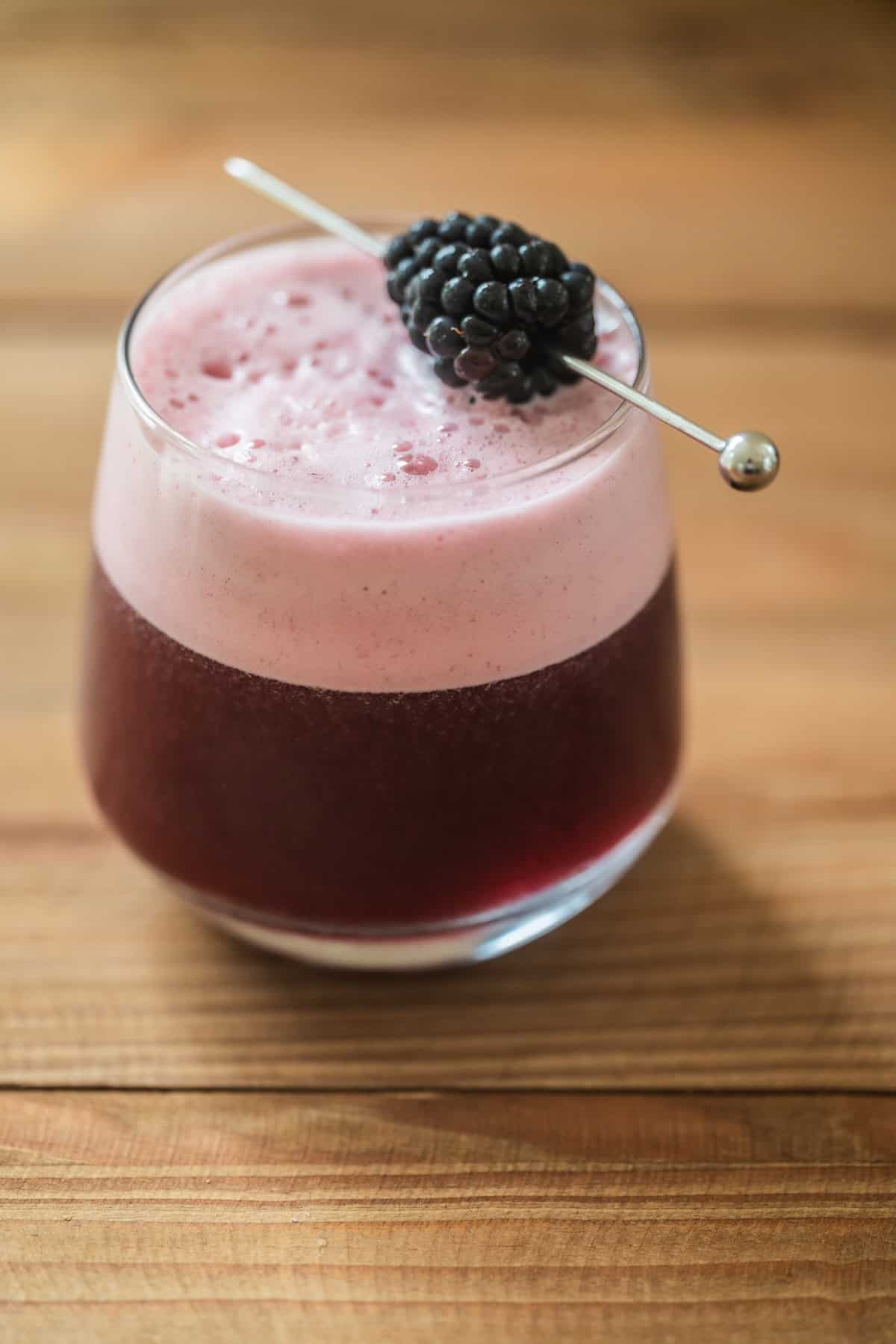 A glass filled with a dark purple drink topped with a layer of pink foam, garnished with a blackberry on a metal pick, sits on a wooden surface.