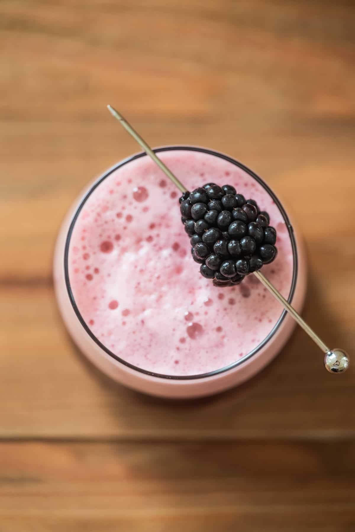 A foamy pink drink in a glass is topped with a skewer holding a fresh blackberry, set on a wooden surface, viewed from above.