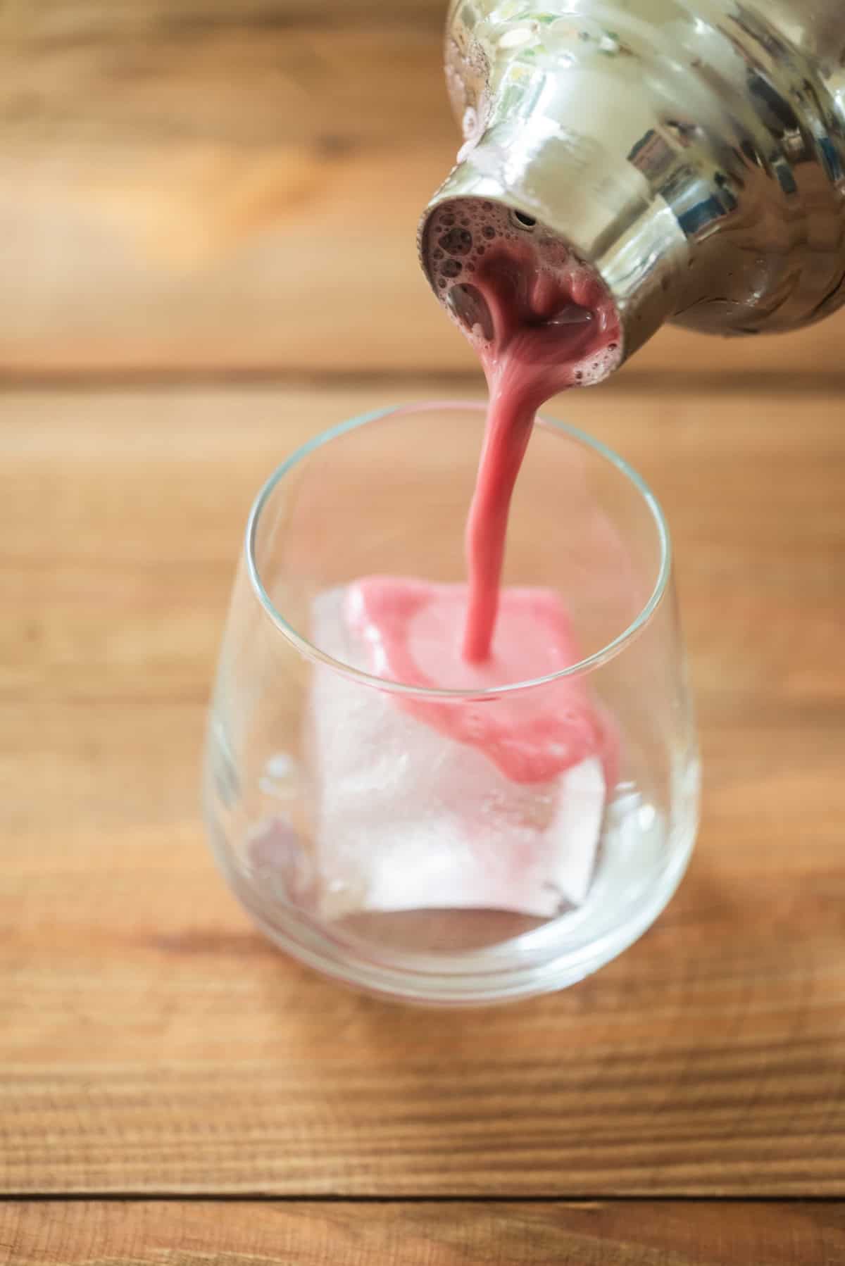 A pink cocktail is being poured from a metal shaker into a glass containing a large ice cube, set on a wooden surface.