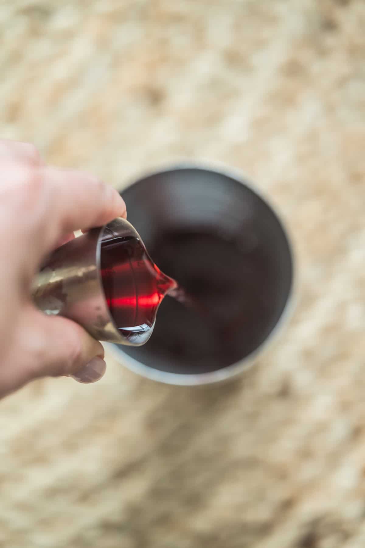 A hand pours a red liquid from a small metal jigger into a metal cup or shaker, set on a textured beige surface.