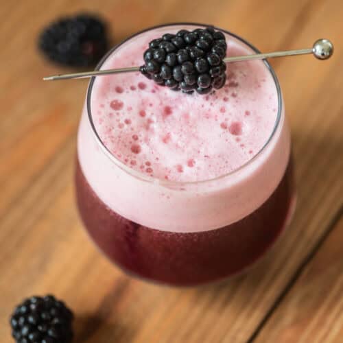 A glass of frothy, pinkish-purple blackberry cocktail garnished with a fresh blackberry on a metal pick, set on a wooden surface with a few loose blackberries nearby.