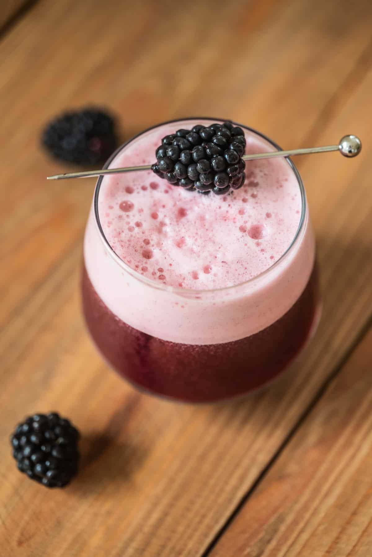 A glass of frothy, pinkish-purple blackberry cocktail garnished with a fresh blackberry on a metal pick, set on a wooden surface with a few loose blackberries nearby.
