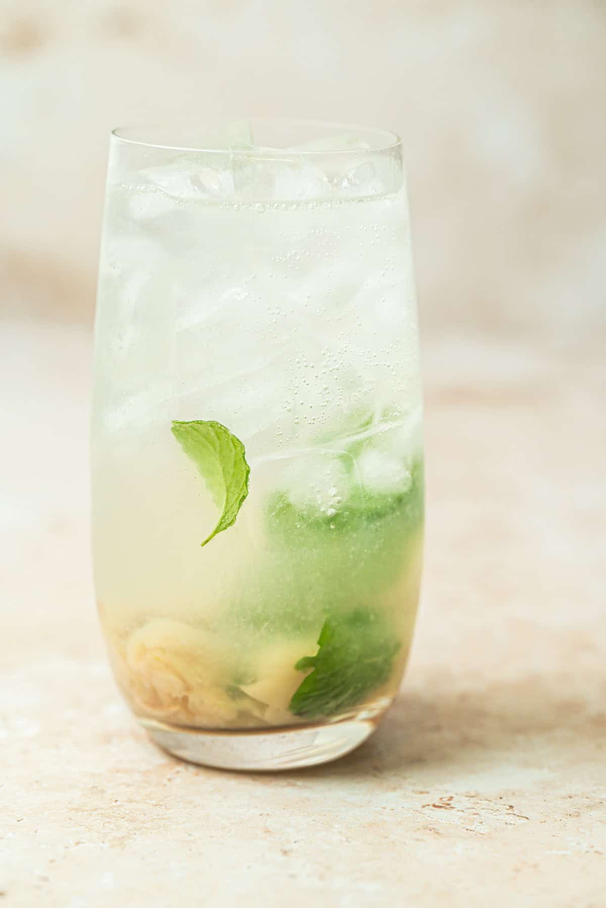 A tall glass filled with ice, a light, bubbly beverage, fresh mint leaves, and lime slices, set on a beige surface with a soft, neutral background.