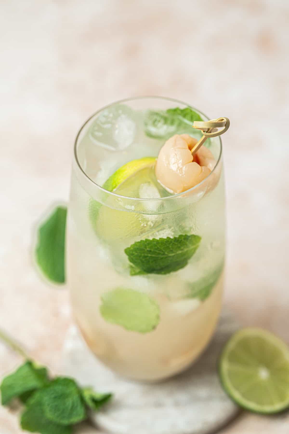 A glass of iced cocktail garnished with fresh mint leaves, lime slices, and a whole lychee on a pick. A lime wedge and mint sprigs rest on the table next to the drink.
