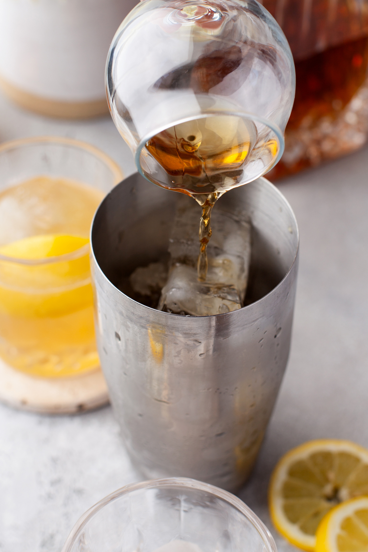 A close-up of whiskey being poured from a glass into a metal cocktail shaker filled with ice, with a cocktail and lemon slices visible in the background.