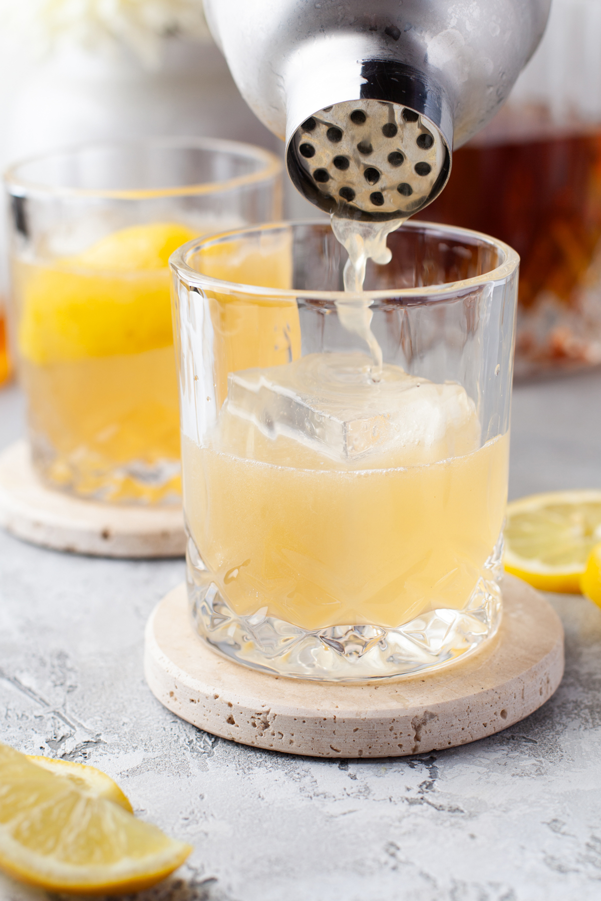 A close-up of a cocktail being poured from a shaker into a short glass with ice, garnished with a lemon twist. Another similar drink sits in the background, and lemon slices rest on the table.