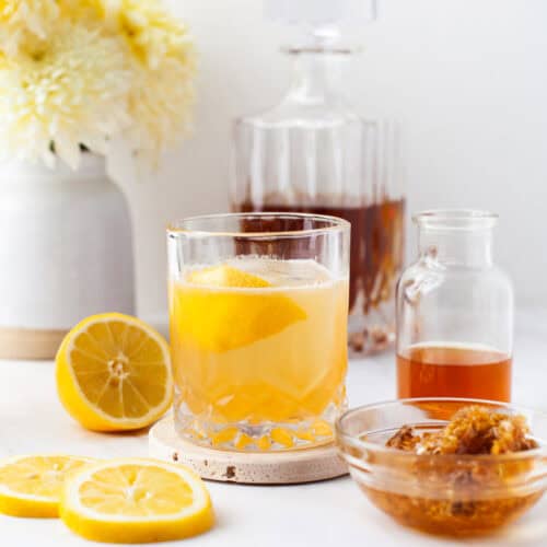 A glass of gold rush cocktail with a lemon twist sits on a coaster, surrounded by lemon slices, a bowl of honey, a small bottle of syrup, and a decanter, with a vase of yellow flowers in the background.