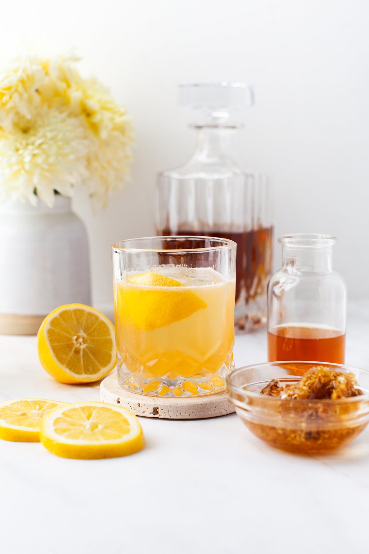 A glass of gold rush cocktail with a lemon twist sits on a coaster, surrounded by lemon slices, a bowl of honey, a small bottle of syrup, and a decanter, with a vase of yellow flowers in the background.