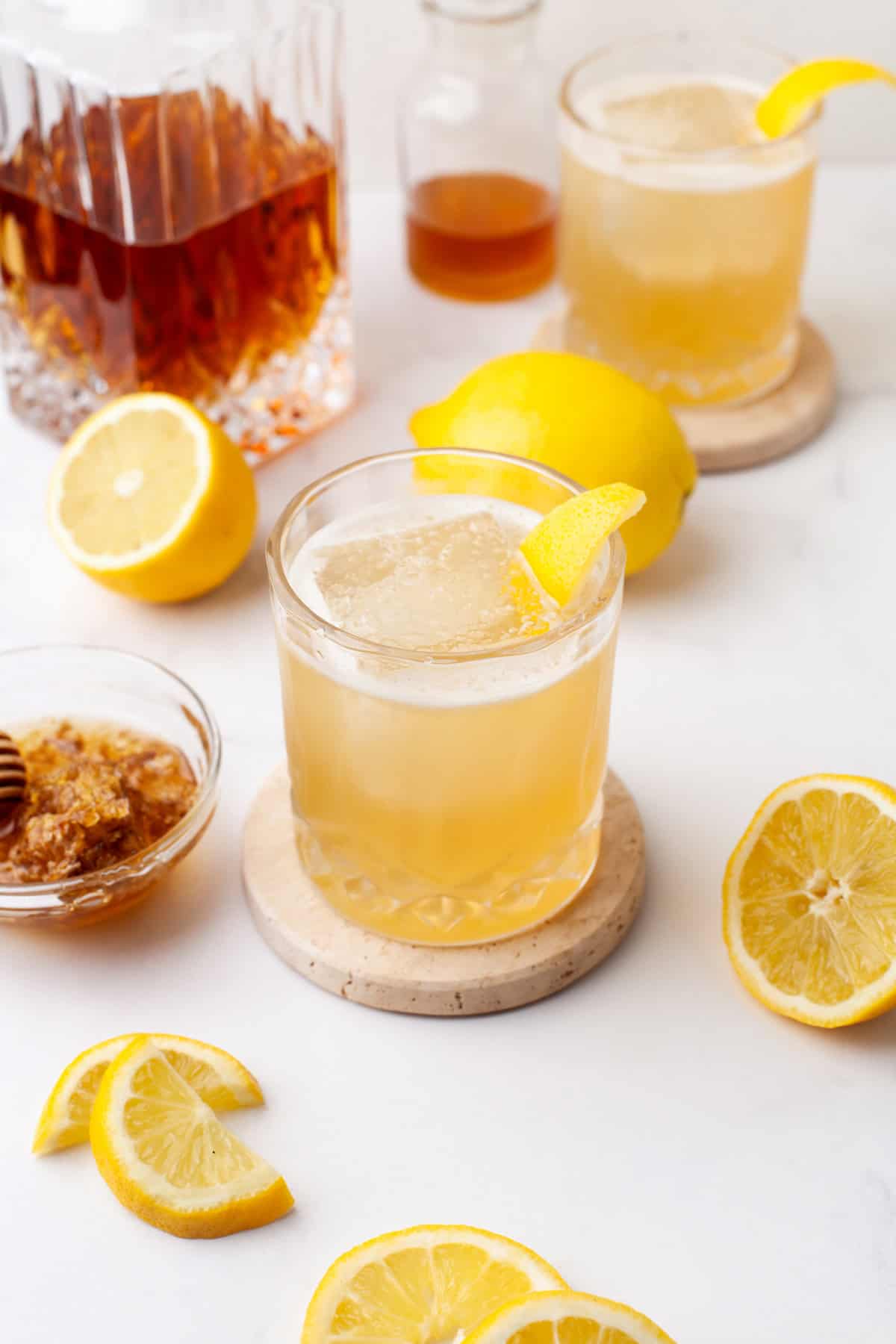 A glass of iced whiskey cocktail garnished with a lemon wedge sits on a coaster, surrounded by sliced lemons, honeycomb in a bowl, a decanter of whiskey, and another drink in the background on a white surface.