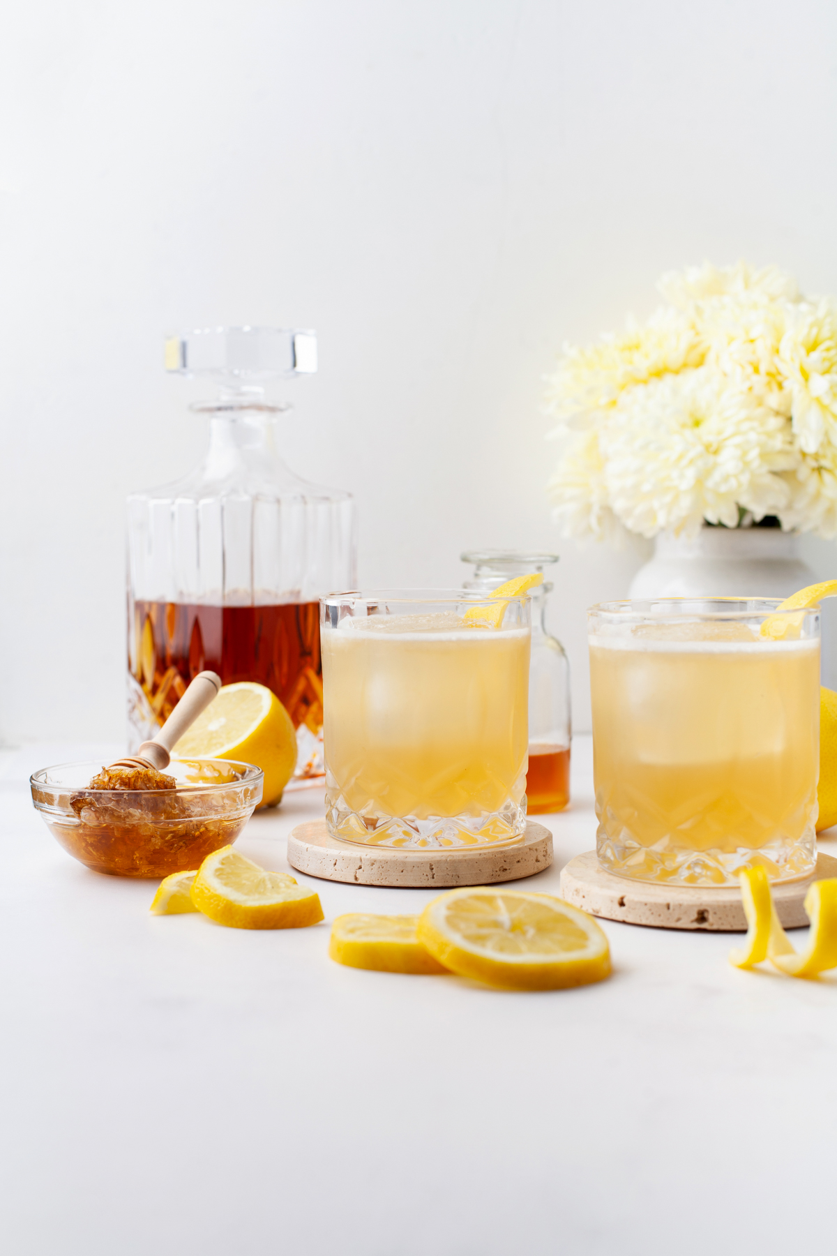 Two glasses of Gold Rush Cocktails with lemon wedges, honey, and a decanter of whiskey on a white surface; white flowers and jars in the background.