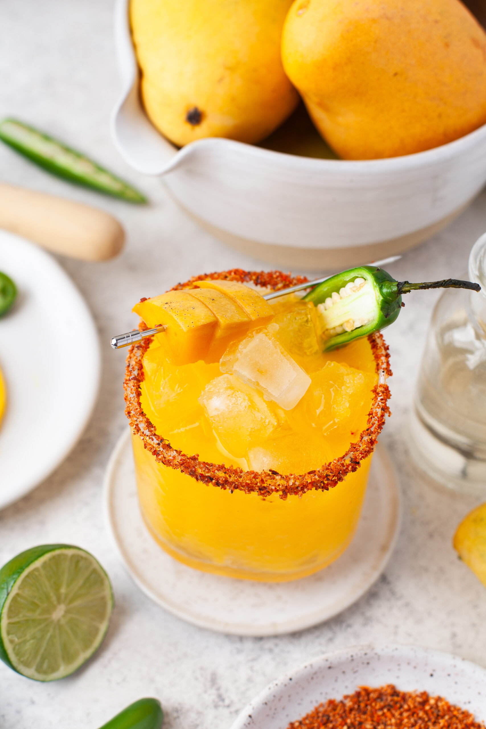 A yellow cocktail with ice sits in a glass rimmed with chili powder, garnished with a mango slice and a jalapeño. Fresh mangoes, lime, and chili powder are nearby on a white surface.