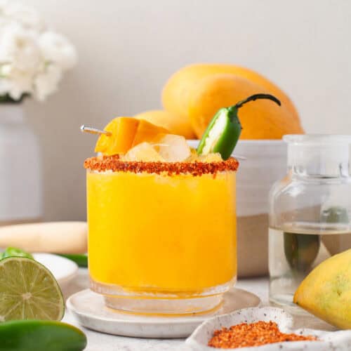 A glass of bright yellow mango chili margarita drink with ice, chili powder rim, mango chunks, and a jalapeño garnish sits on a plate. Fresh mango, lime, and a jar of clear liquid are nearby. Flowers and fruit decorate the background.