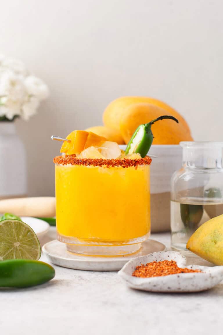 A glass of bright yellow mango chili margarita drink with ice, chili powder rim, mango chunks, and a jalapeño garnish sits on a plate. Fresh mango, lime, and a jar of clear liquid are nearby. Flowers and fruit decorate the background.