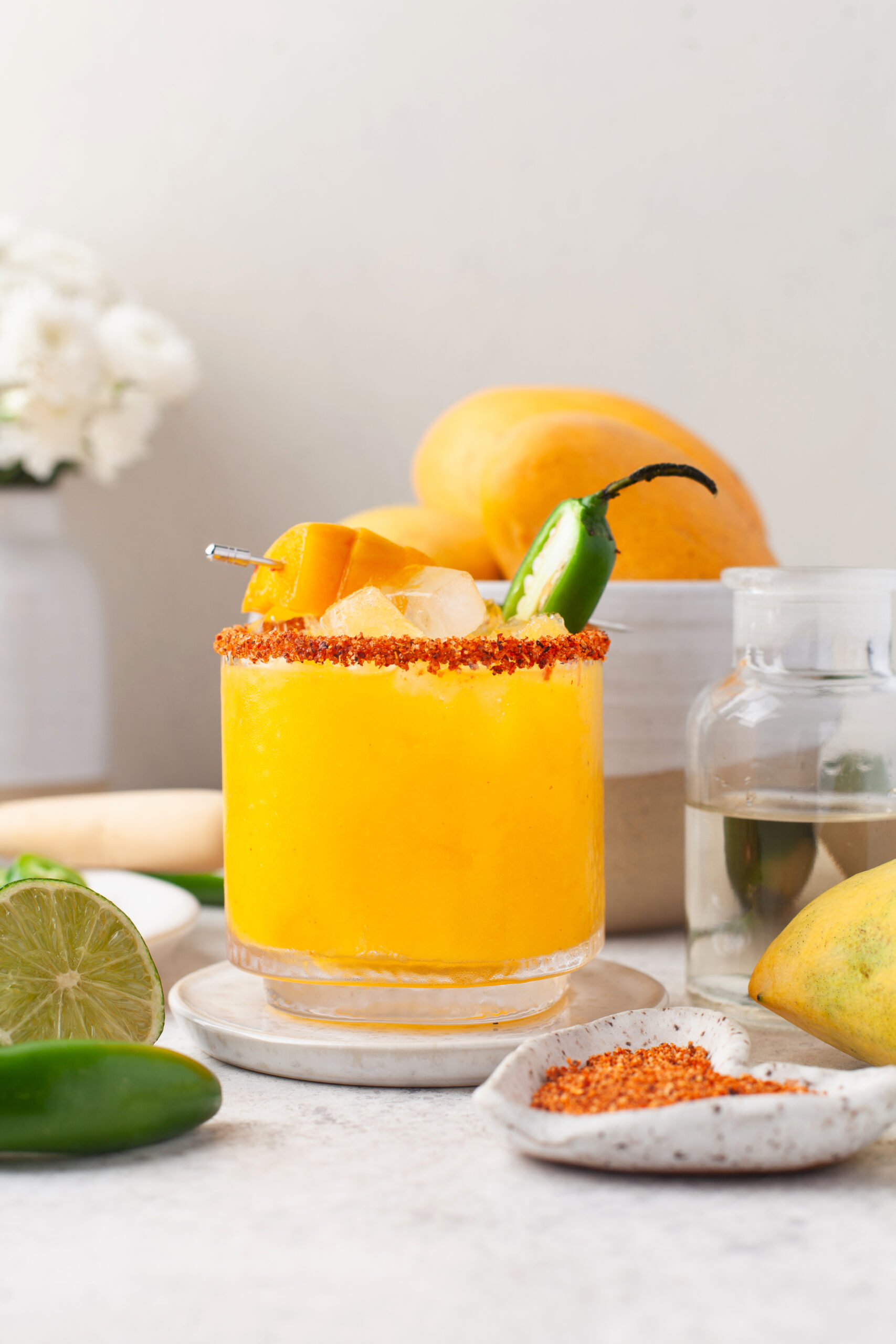 A glass of bright yellow mango drink with ice, chili powder rim, mango chunks, and a jalapeño garnish sits on a plate. Fresh mango, lime, and a jar of clear liquid are nearby. Flowers and fruit decorate the background.