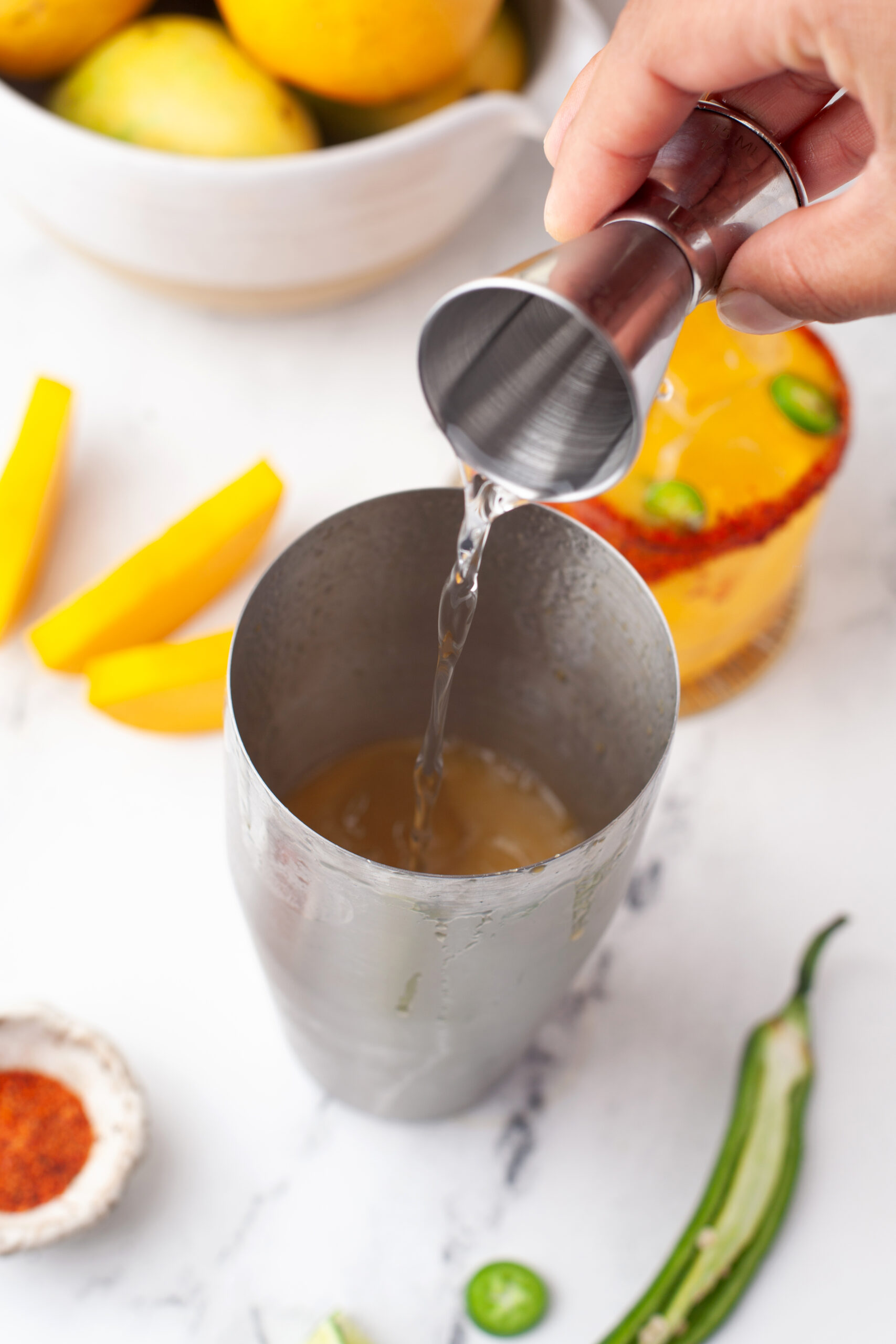 Hand pouring liquid from a jigger into a metal cocktail shaker, with sliced fruit, a drink, and a bowl of lemons nearby on a white surface.