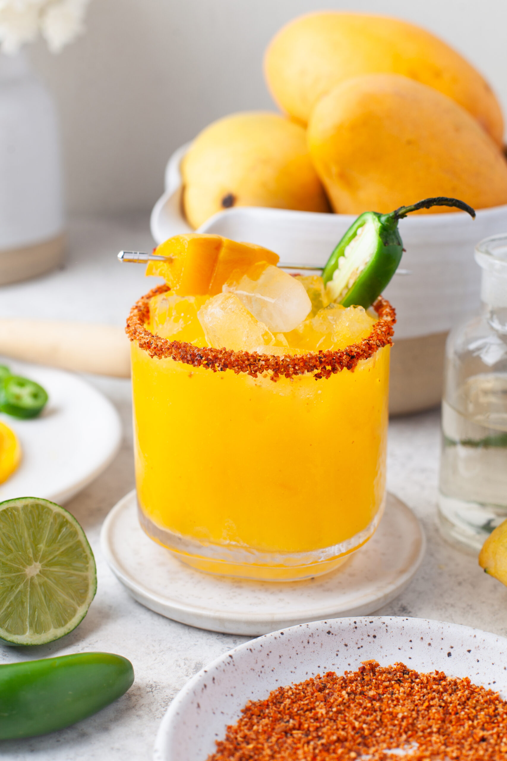 A glass of mango cocktail with chili powder rim, ice, mango cubes, and a jalapeño slice garnish, surrounded by fresh mangoes, lime, and a bowl of chili powder on a white surface.