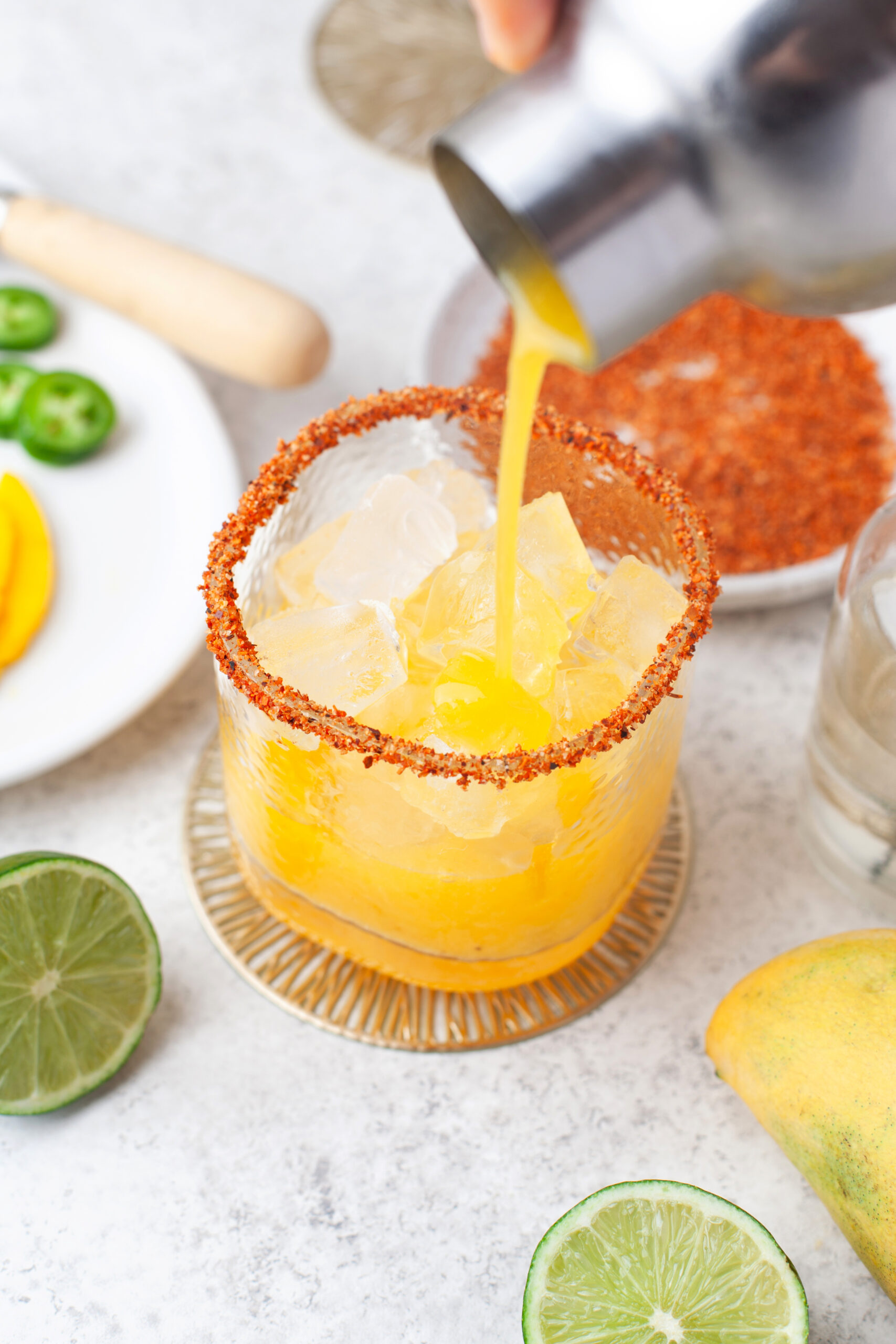 A yellow cocktail with ice is poured into a glass rimmed with chili powder. Lime, sliced jalapeño, mango, and chili powder are nearby on a white surface.