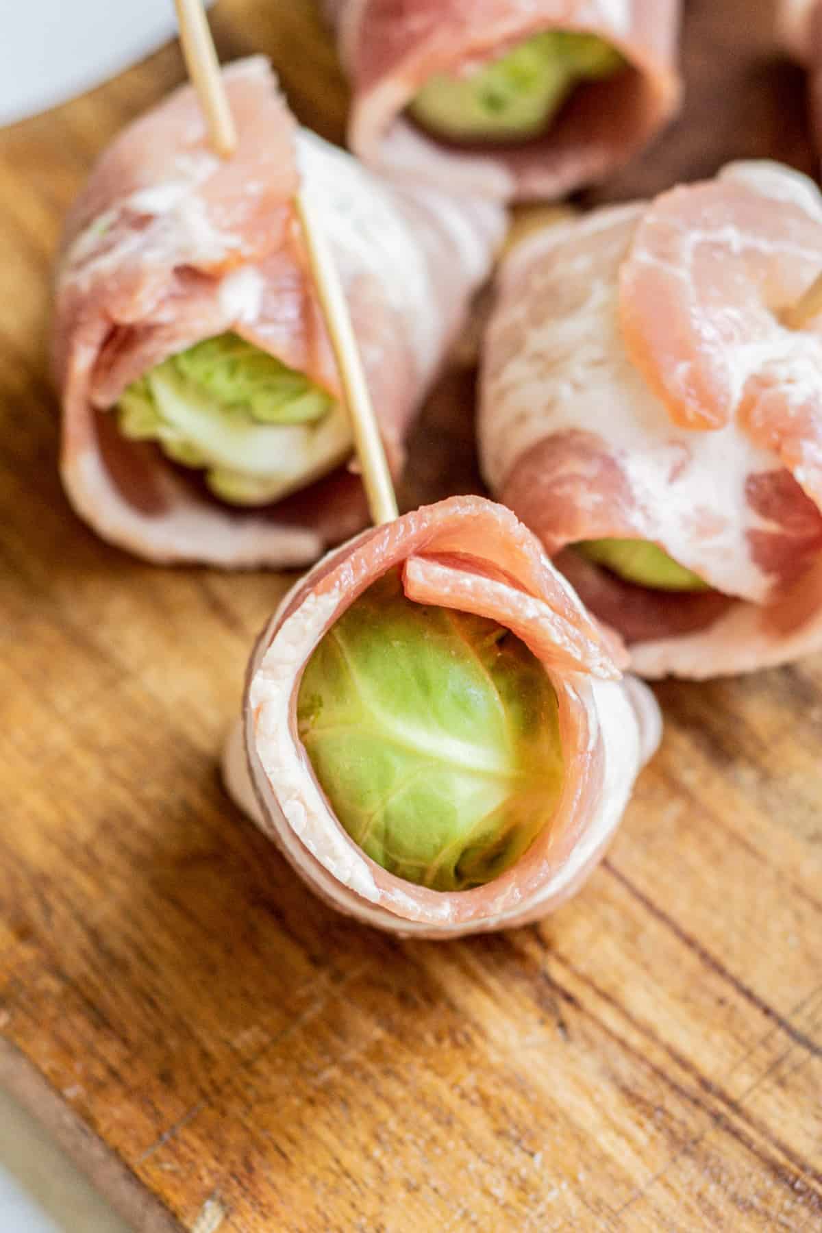 Close-up of Brussels sprouts wrapped in raw bacon and secured with wooden toothpicks, arranged on a wooden cutting board.