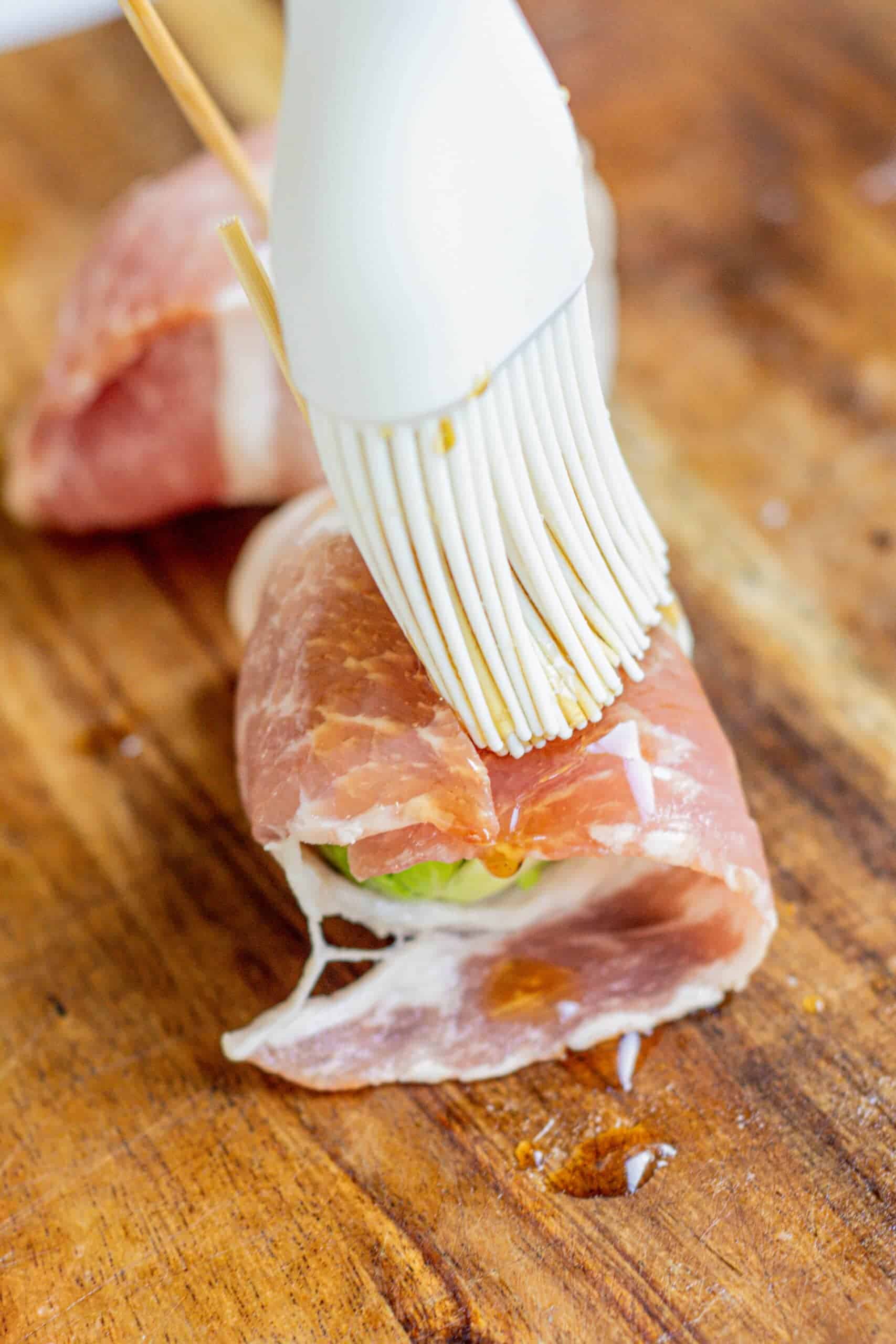 A silicone brush coats a prosciutto-wrapped appetizer with oil or glaze on a wooden cutting board.