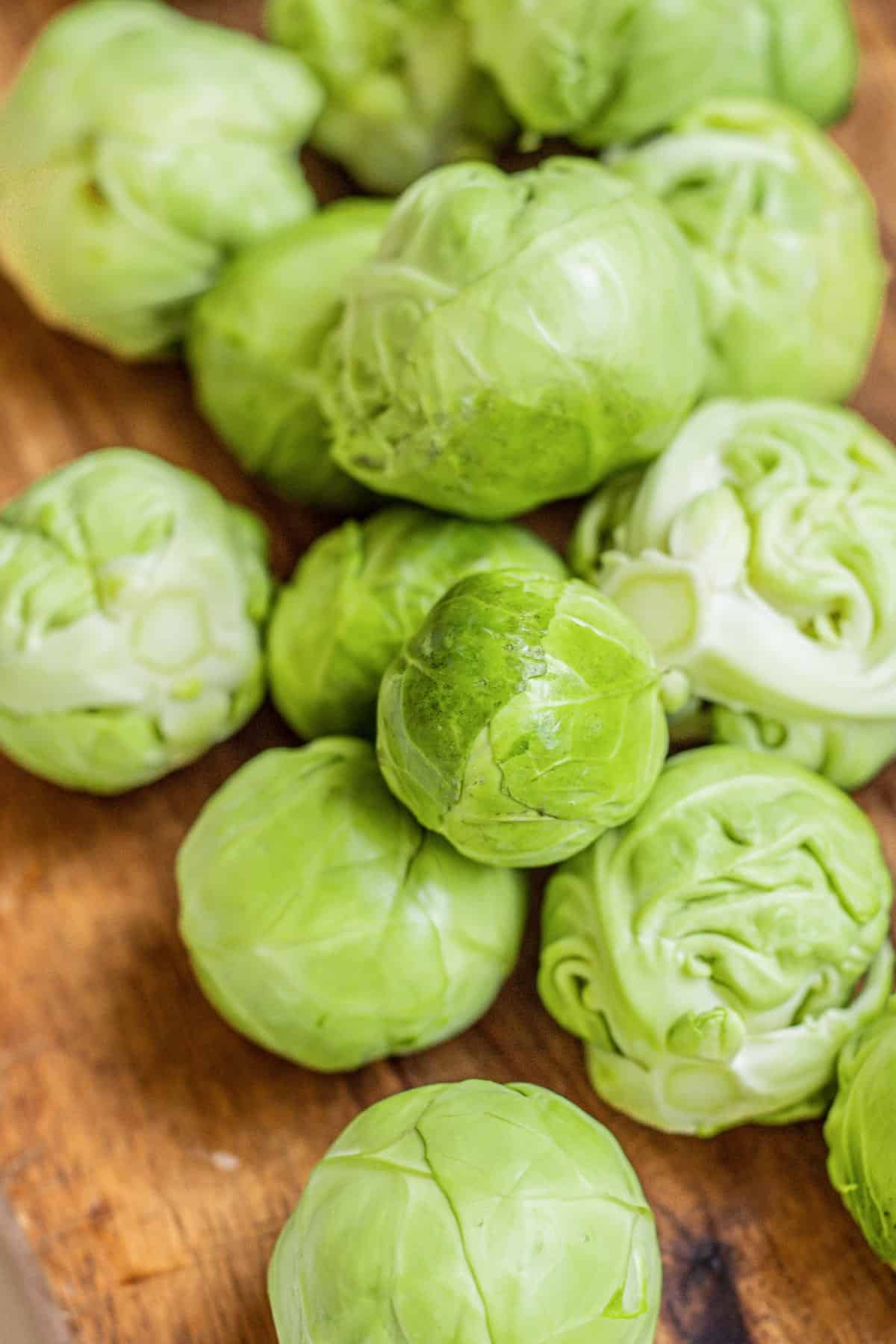 Several fresh Brussels sprouts are piled on a wooden surface, showing their vibrant green color and tightly packed leaves.