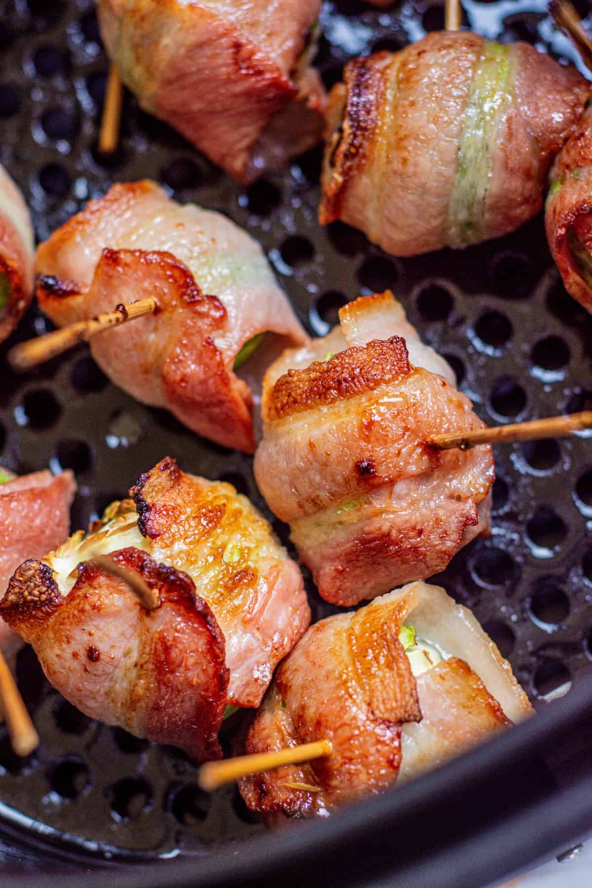 Several bacon-wrapped jalapeño poppers secured with toothpicks are arranged in an air fryer basket, showing crispy, golden-brown edges.