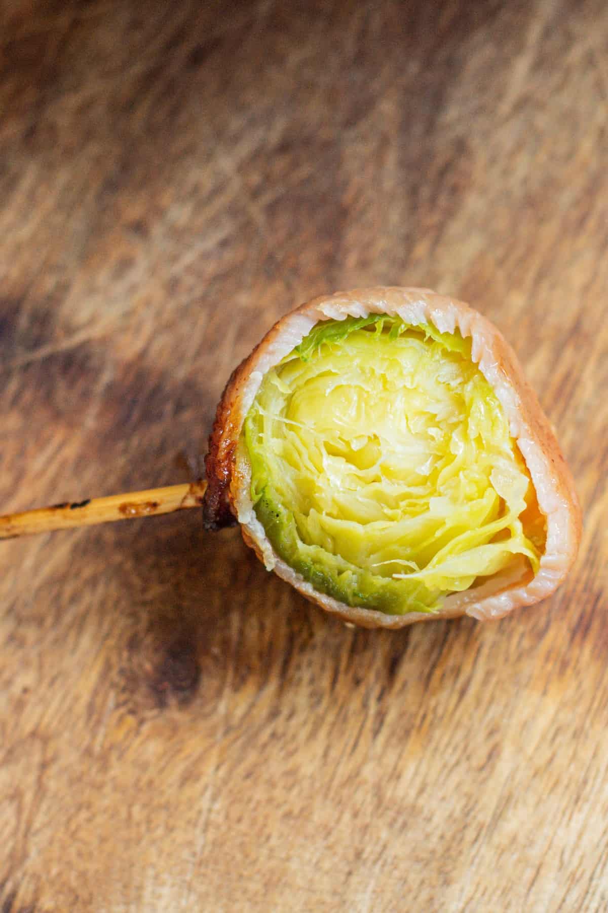 A Brussels sprout wrapped in bacon on a wooden skewer sits on a rustic wooden surface.