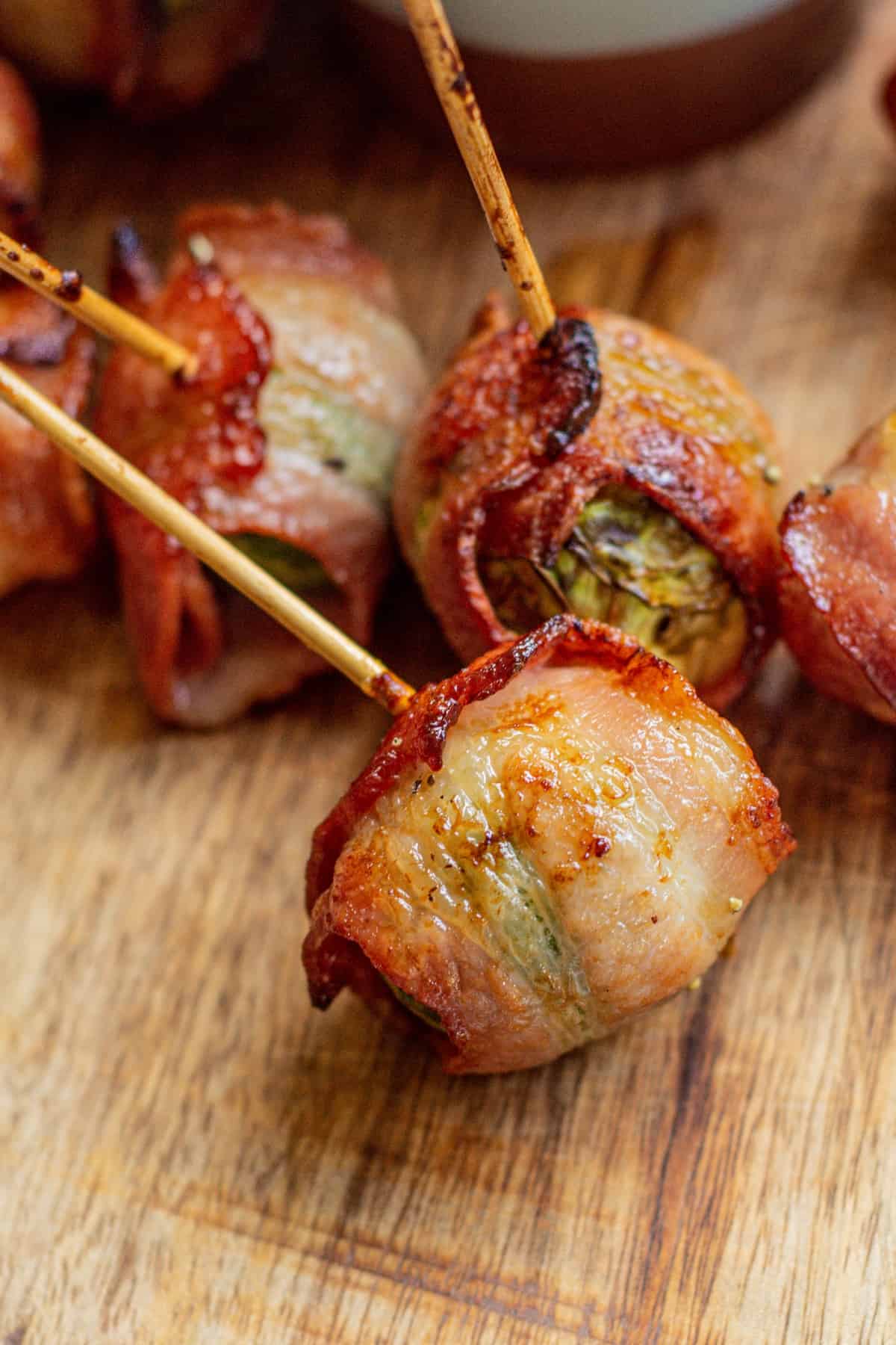 Three bacon-wrapped Brussels sprouts skewered with wooden sticks rest on a wooden surface. The bacon is crispy and browned, and the Brussels sprouts are visible inside.