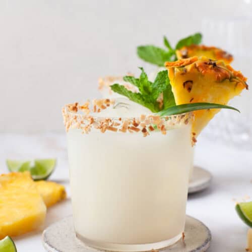 A frosty cocktail in a glass with a toasted coconut rim, garnished with a pineapple wedge, mint, and a slice of lime. Pineapple and lime pieces are on the white surface nearby.