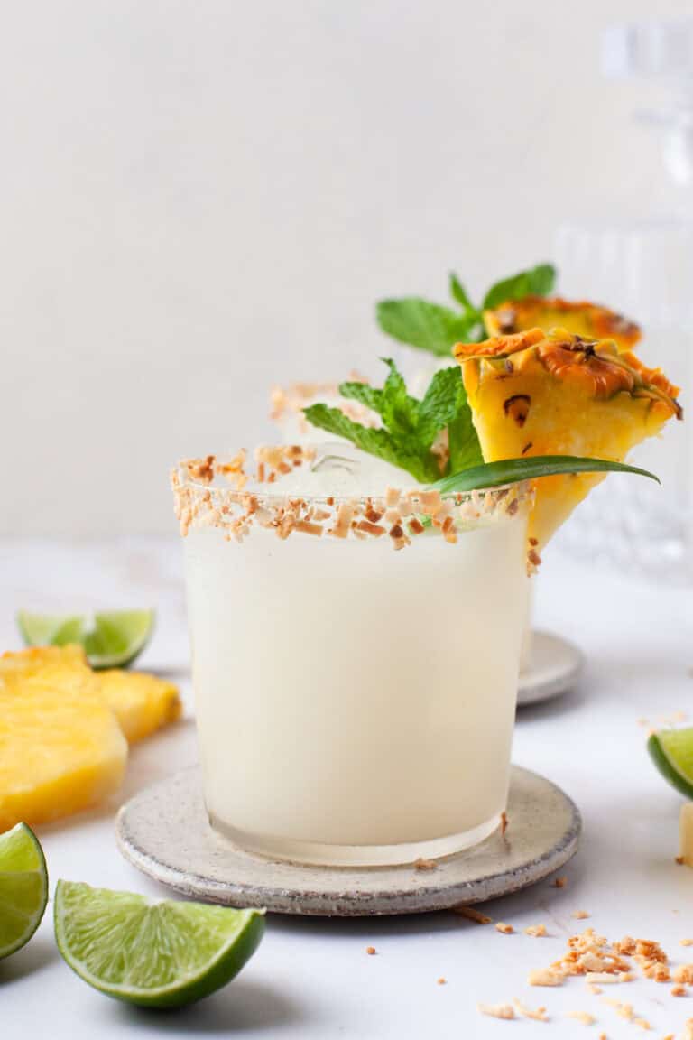 A frosty cocktail in a glass with a toasted coconut rim, garnished with a pineapple wedge, mint, and a slice of lime. Pineapple and lime pieces are on the white surface nearby.