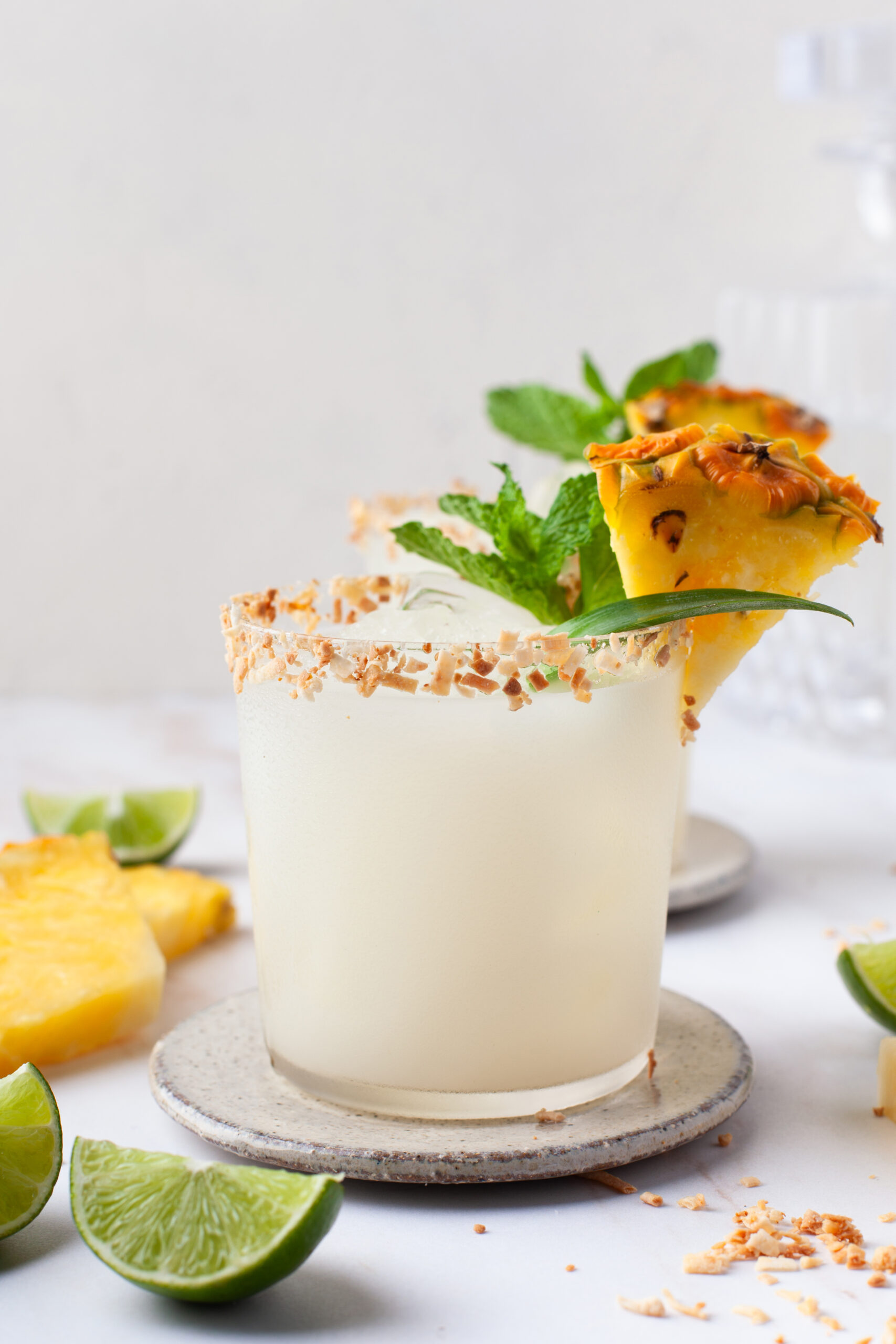 A frosty cocktail in a glass with a toasted coconut rim, garnished with a pineapple wedge, mint, and a slice of lime. Pineapple and lime pieces are on the white surface nearby.