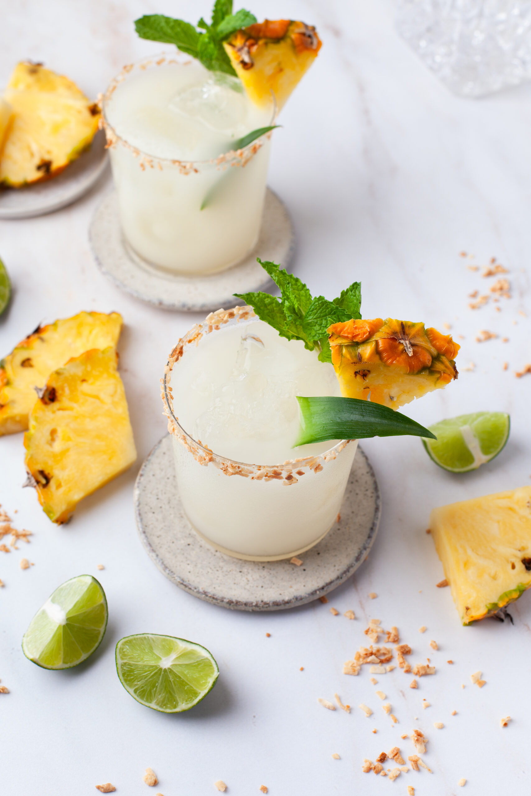 Two tropical drinks with ice, garnished with pineapple, mint, and lime, sit on coasters. Pineapple slices, toasted coconut, and lime wedges are scattered on a marble surface nearby.