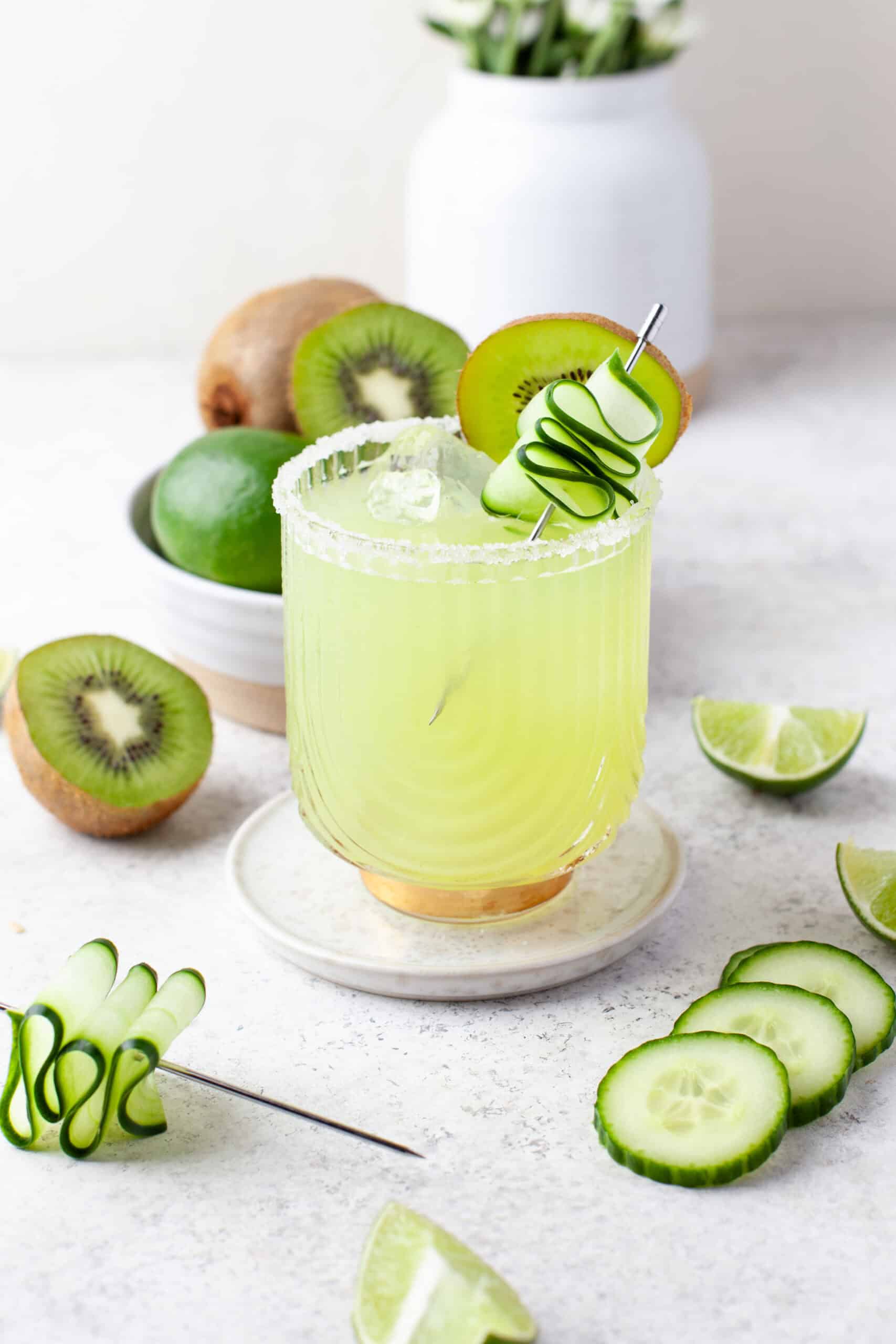 A green cocktail garnished with a cucumber ribbon and kiwi slice, surrounded by fresh kiwi, lime, cucumber slices, and a bowl of fruit on a light surface.