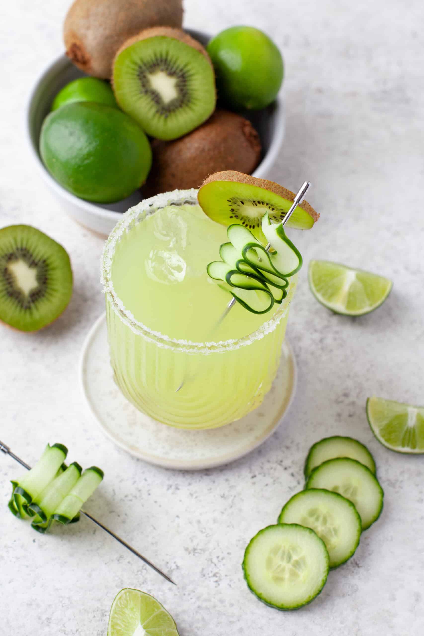 A green cocktail garnished with a kiwi slice and twisted cucumber sits on a coaster, surrounded by sliced cucumber, lime wedges, and a bowl of whole kiwi and limes.