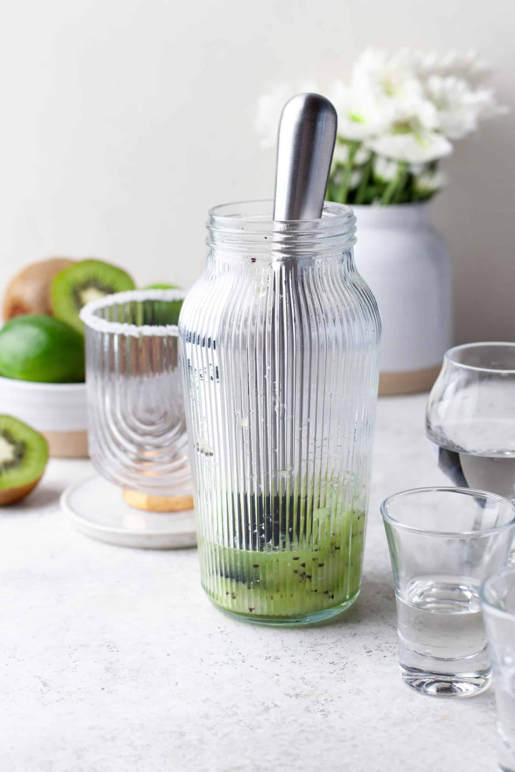 A glass jar with mashed kiwi and a muddler inside sits on a white table, surrounded by whole and sliced kiwis, a glass of water, an empty glass, and a vase of white flowers in the background.