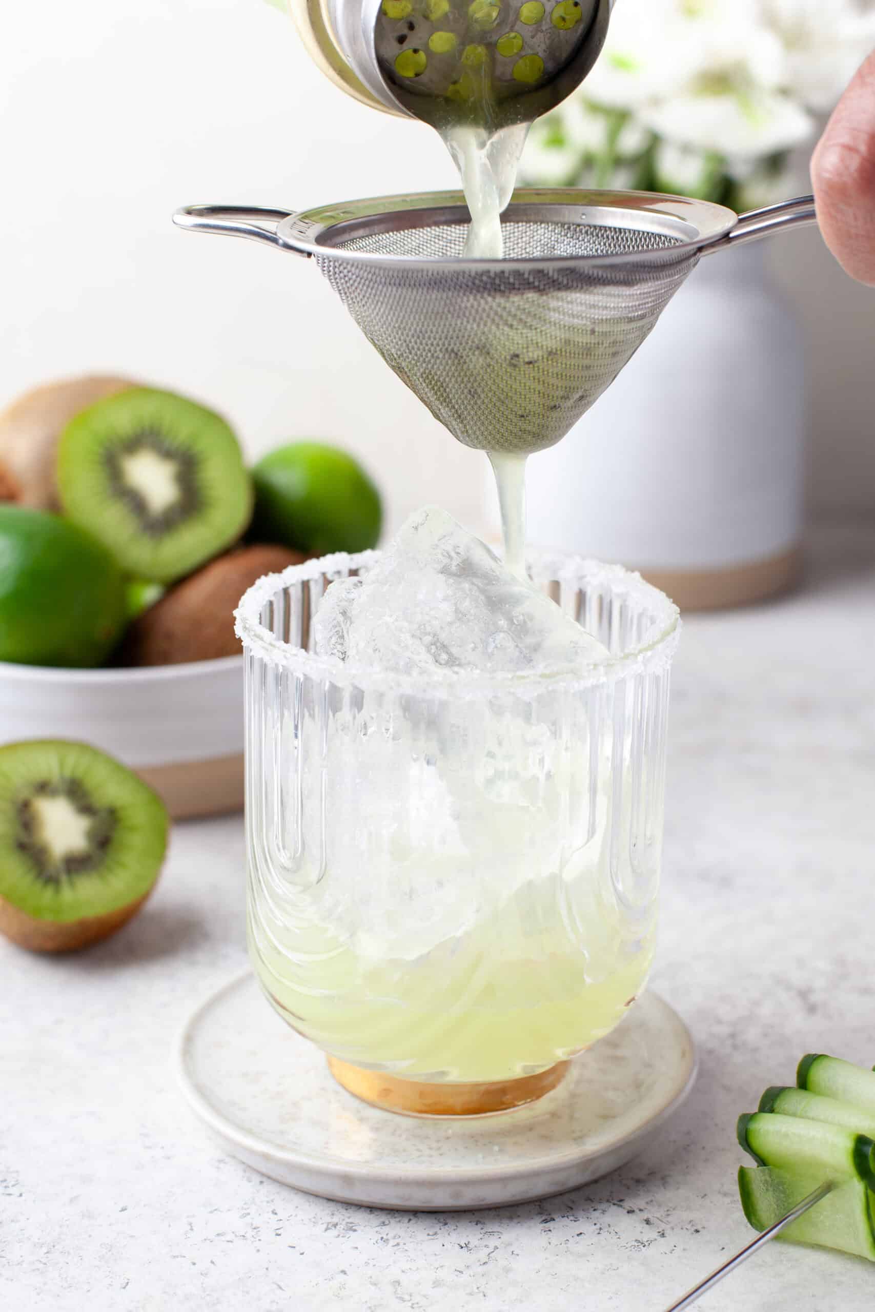Kiwi juice is being poured through a strainer into a glass with ice, set on a small plate. Fresh kiwi and limes are in the background, and the glass rim is coated with salt or sugar.