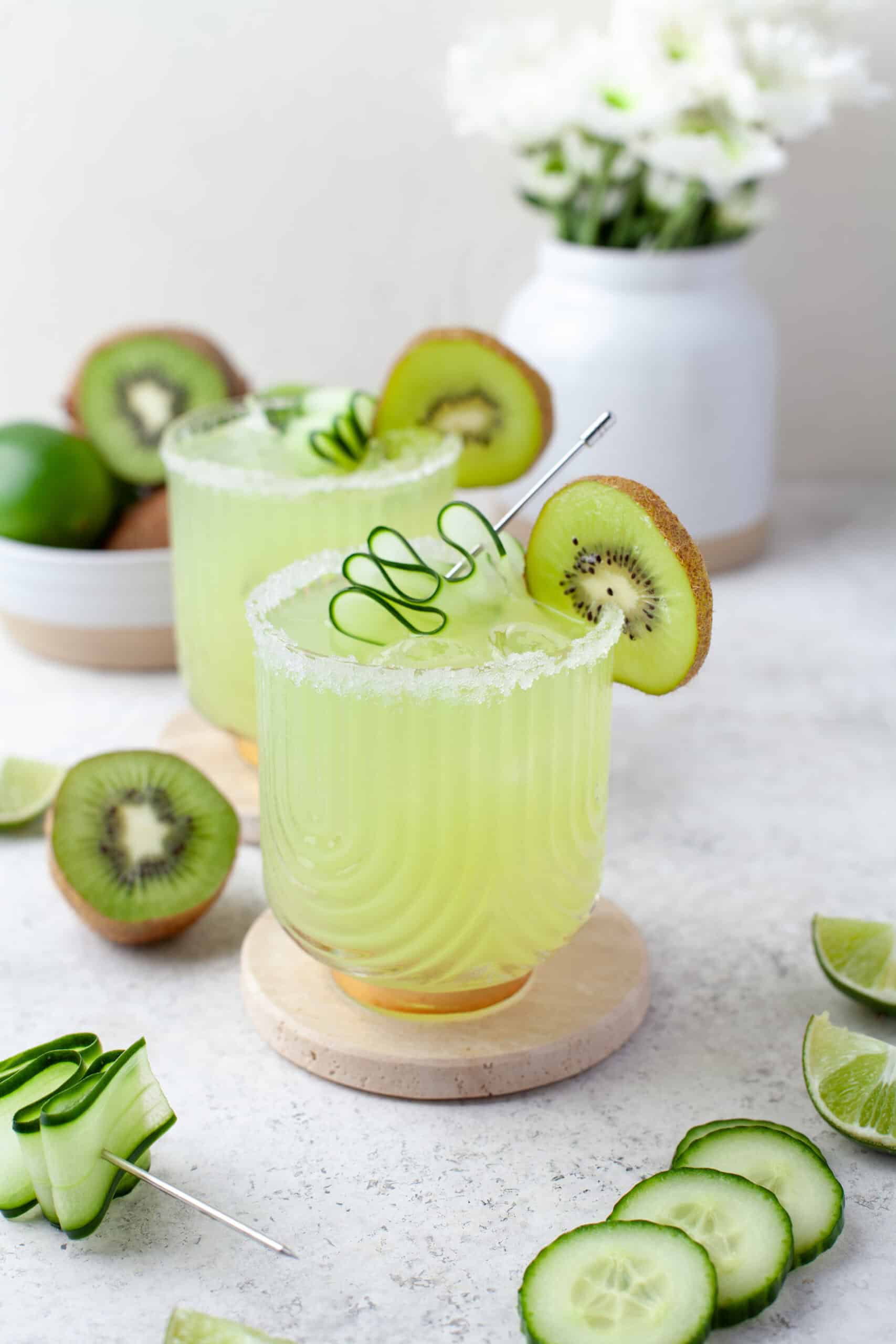 Two green cocktails garnished with kiwi slices and cucumber spirals sit on coasters. Fresh kiwis, cucumbers, lime wedges, and a bowl of limes are nearby. White flowers in a vase are blurred in the background.