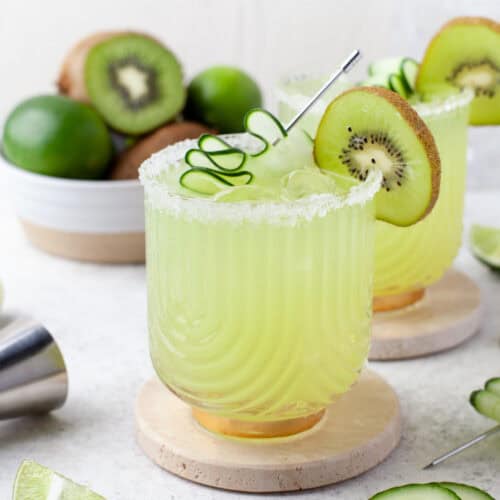 A glass of green cocktail with ice, garnished with a cucumber spiral and a kiwi slice. Sliced cucumber, lime wedges, and a bowl of kiwis and limes are in the background. The drink has a salted rim.