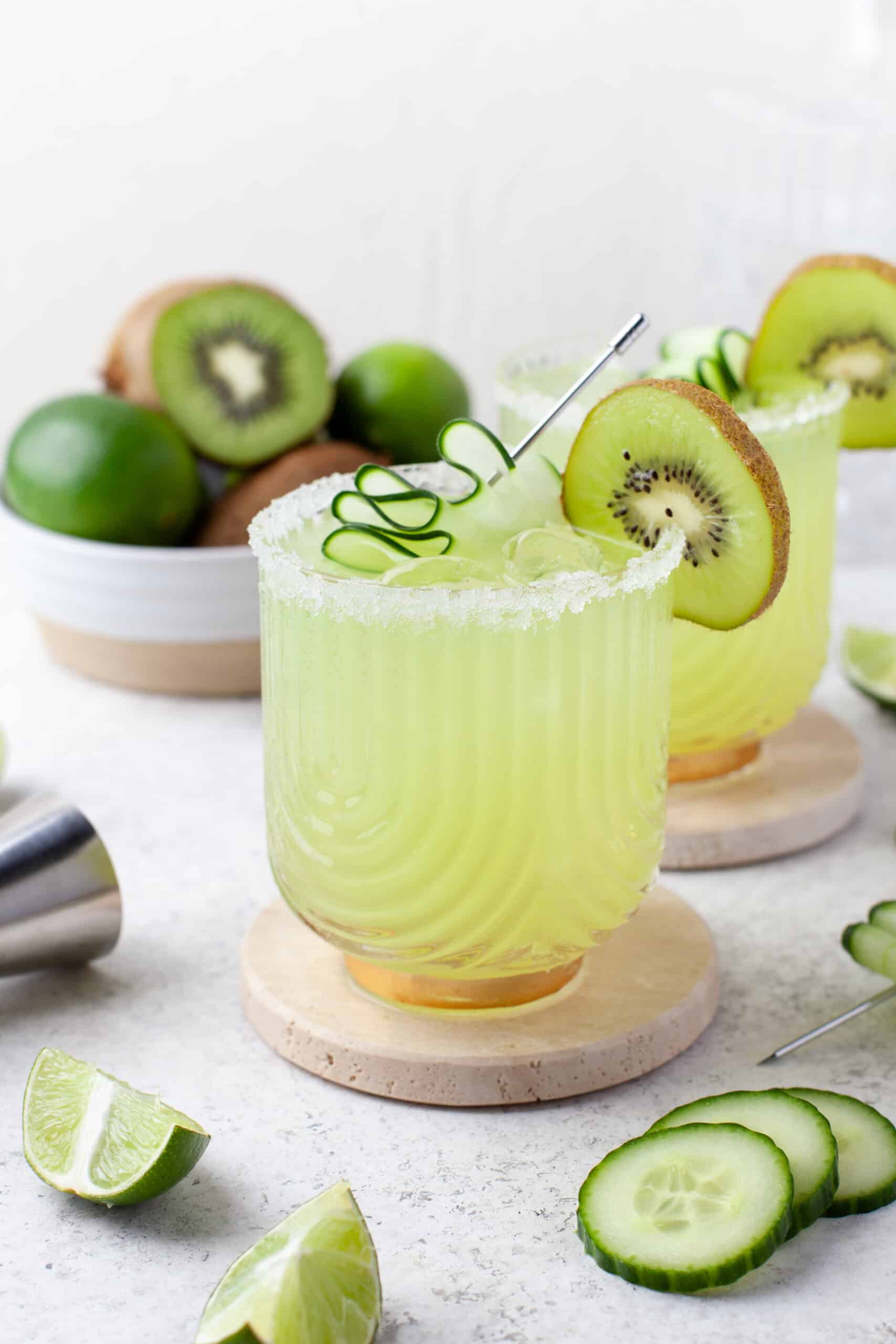 A glass of green cocktail with ice, garnished with a cucumber spiral and a kiwi slice. Sliced cucumber, lime wedges, and a bowl of kiwis and limes are in the background. The drink has a salted rim.