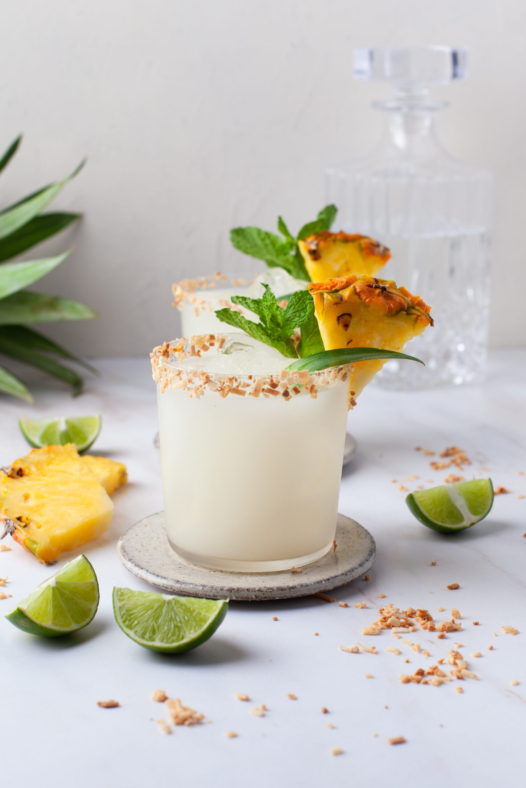 Two cocktails in glasses with toasted coconut rims, garnished with pineapple, mint, and pineapple leaves. Lime wedges and pineapple slices are scattered on a white surface. A decanter and pineapple top are in the background.