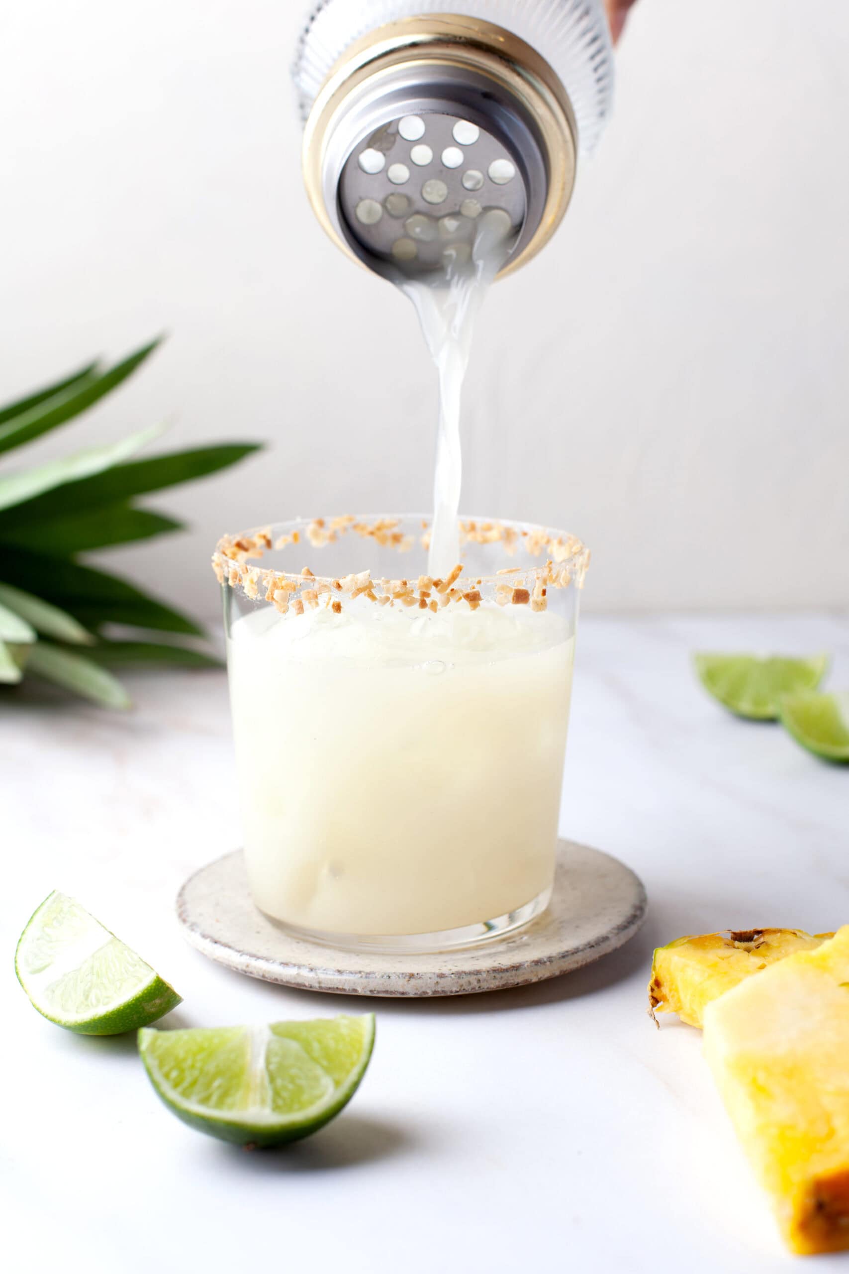 A glass with a toasted rim is being filled with a light-colored drink from a shaker. Lime wedges and pineapple pieces are on the white surface nearby.