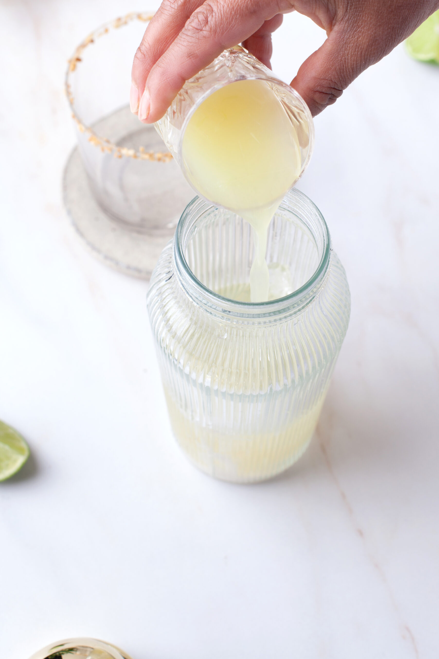 A hand pours yellow juice from a small glass into a ribbed glass jar. A lime and a salt-rimmed glass are visible on a white surface in the background.