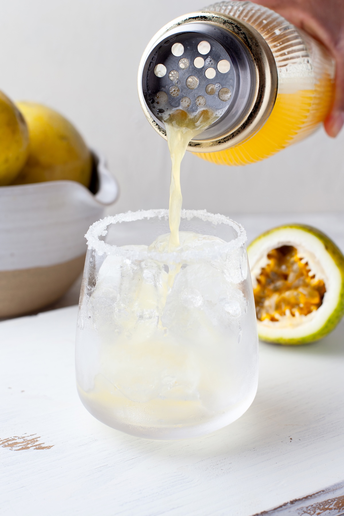 A hand pours yellow juice from a cocktail shaker into an ice-filled glass with a salted rim; a halved passion fruit and a bowl of yellow fruits are in the background.