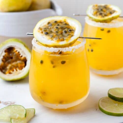 Two glasses of yellow passion fruit cocktail with salted rims, garnished with passion fruit halves, sit on a white board next to lime slices. A bowl and halved passion fruit are in the background.