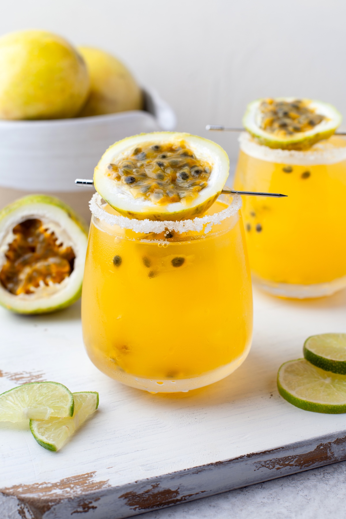 Two glasses of yellow passion fruit cocktail with salted rims, garnished with passion fruit halves, sit on a white board next to lime slices. A bowl and halved passion fruit are in the background.