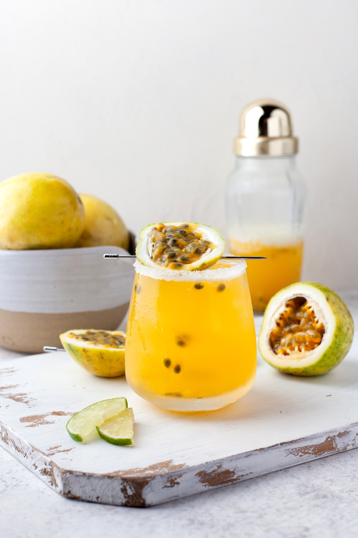 A glass of yellow passion fruit juice with ice, garnished with half a passion fruit. A bowl of passion fruits, lime wedges, and a cocktail shaker are in the background on a white surface.
